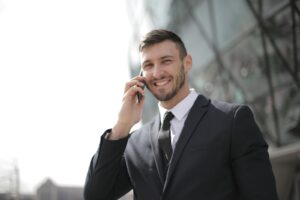 Un homme d'affaires souriant en costume passe un appel téléphonique à l'extérieur d'un bâtiment moderne en verre.